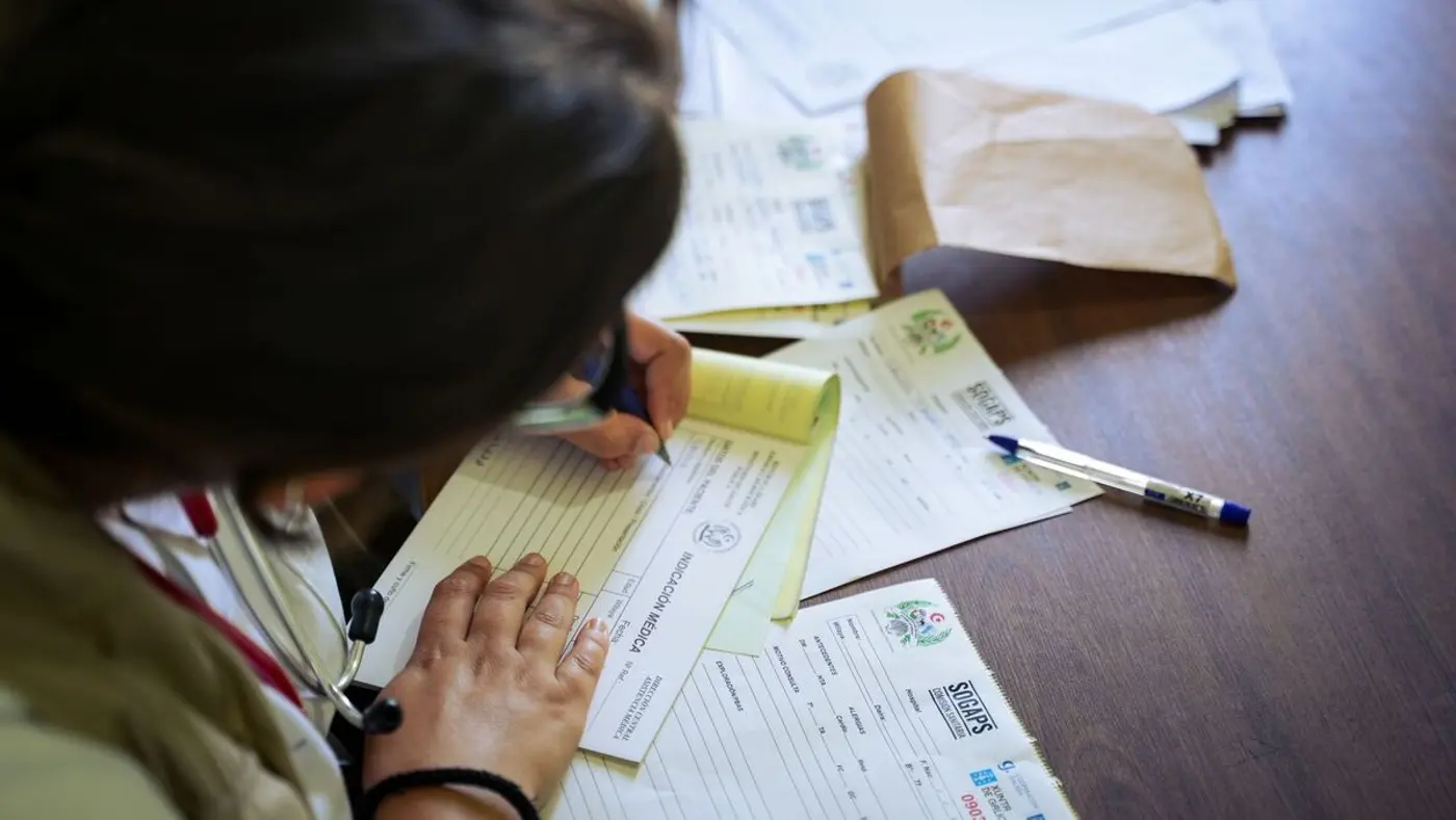 A clinician fills out paper medical forms at a wooden desk